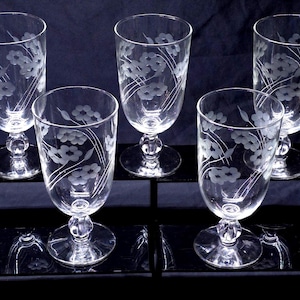 May include: A set of five clear glass goblets, each featuring a delicate etched floral pattern with thin stems and small blossoms. The glasses have a rounded bowl, a short stem with a decorative spherical node, and a circular base for stability.