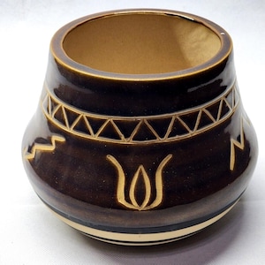 May include: A dark brown ceramic pot with a wide, rounded body and a circular opening. The exterior features etched light-colored geometric patterns, including a band of triangles near the rim and stylized floral motifs around the center.