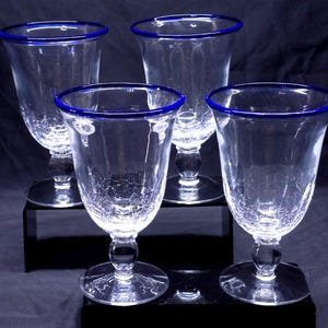 May include: A set of four clear glass goblets featuring a distinct crackle texture throughout the bowl and a vibrant cobalt blue rim. Each goblet is supported by a clear glass stem with a spherical accent and a circular base for stability.
