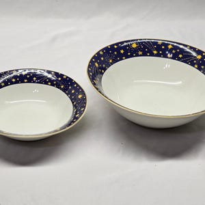 May include: Two round ceramic bowls with a white interior and a wide dark blue rim featuring a pattern of yellow stars and shooting stars. The bowls have a thin gold-colored trim along the top edge, with one bowl being noticeably larger than the other.