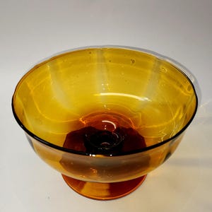 Peut inclure: Un bol en verre ambré translucide avec une large ouverture circulaire et un bord festonné. Le bol présente une base en verre brun foncé profond qui se transforme en une teinte jaune doré plus claire vers le haut, reposant sur un pied.