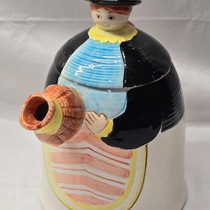 May include: A ceramic teapot shaped like a person wearing a black hat, a black jacket, a blue striped apron, and a pink striped skirt. The spout is designed to look like a woven basket held by the figure, featuring a brown and tan texture.