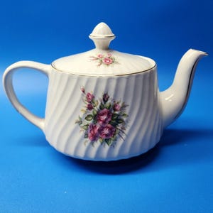 Puede incluir: Una tetera de porcelana blanca con una textura acanalada en espiral y un delicado patrón floral de rosas rosas y moradas. La tetera incluye una tapa a juego con un pequeño motivo de rosa y un borde dorado en la abertura, el asa y el pico.