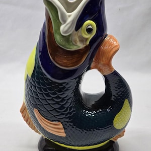 May include: A ceramic jug shaped like a fish with a wide open mouth serving as the spout. The body is dark blue with textured scales, featuring yellow patches, orange fins, and a green interior. It stands on a solid black circular base.