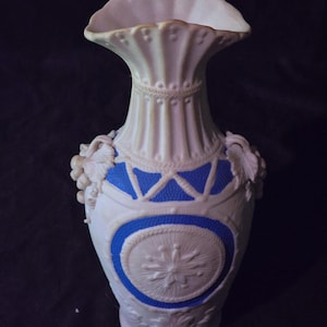 May include: A ceramic vase featuring a fluted neck and a scalloped rim. The body is decorated with white relief patterns against a vibrant blue background, including circular medallions, geometric lines, and sculpted grape clusters on the sides.
