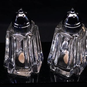 May include: A pair of clear glass salt and pepper shakers with a faceted, geometric design and polished chrome dome-shaped lids. Each shaker features a wide base that tapers toward the top, with vertical panels creating a distinct angular profile.