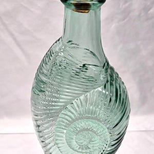 May include: A light green glass bottle with a textured, spiralling shell-like pattern on its surface. The bottle features a narrow neck and is sealed with a cylindrical cork stopper. The glass is translucent, showing subtle variations in colour.
