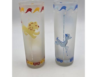 May include: Two tall frosted glass tumblers featuring carousel-themed graphics. The left glass displays a yellow lion with red and yellow scalloped trim. The right glass features a blue deer with light blue scalloped trim and vertical pole details.