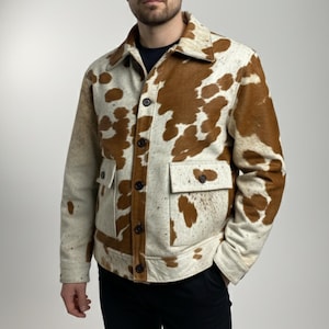 Könnte beinhalten: Eine Jacke mit Knöpfen und Kuhfellmuster. Die Jacke hat eine cremefarbene Basis mit braunen Flecken, zwei Vordertaschen und einen Kragen. Sie hat dunkelbraune Knöpfe und eine klassische Passform.