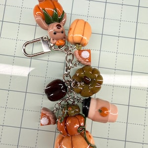 Op de afbeelding: Een zilverkleurige sleutelhanger met diverse miniatuurbedels, waaronder een beer in een pompoenkostuum, verschillende oranje en groene pompoenen, twee stukjes pompoentaart met slagroom, een donkere bes en een koffiebeker met pompoenicoon.