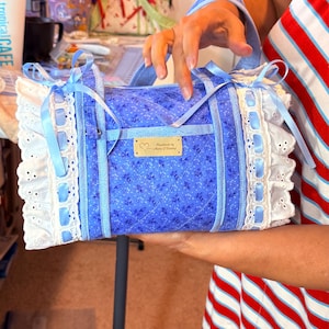 Peut inclure: Un sac en tissu bleu matelassé avec un petit motif floral, agrémenté d'une bordure en dentelle blanche à œillets et de nœuds en ruban bleu clair. Une étiquette rectangulaire en métal doré est centrée sur le devant avec le texte "Handmade by Marie J Banting".