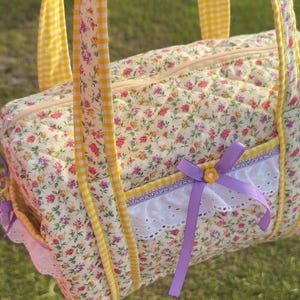 May include: A floral print quilted bag with yellow gingham trim and handles. The bag features a white lace trim, a purple ribbon, and a small yellow flower accent. The bag has a zipper closure.