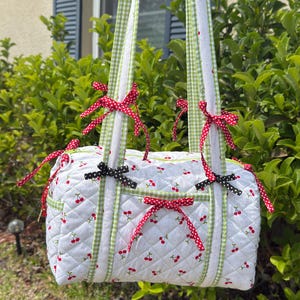 May include: A white quilted handbag with a cherry print, green gingham trim, and red polka dot bows. The bag has two handles and is decorated with black and red bows. The bag is hanging outside in front of green foliage.