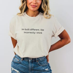 Könnte beinhalten: Cremefarbenes T-Shirt mit dem schwarzen Text "im built different. like incorrectly i think". Das T-Shirt wird mit einer blauen Jeans kombiniert. Die Ärmel sind hochgekrempelt.