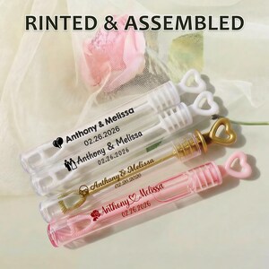May include: Clear plastic bubble wands with heart-shaped handles. Each wand is personalized with the names "Anthony & Melissa" and the date "02.26.2026". The text "RINTED & ASSEMBLED" is at the top. Some wands have gold accents.