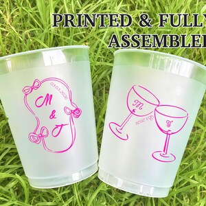 May include: Two frosted plastic cups with pink designs and text. One cup features the letters "M & J" with a date, and the other has two wine glasses with the letters "M & J" and a date. The text "PRINTED & FULLY ASSEMBLED" is at the top.