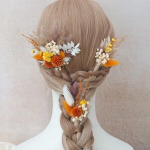 Könnte beinhalten: Geflochtene Frisur mit drei floralen Haarspangen. Die Spangen zeigen getrocknete Blumen in Orange, Gelb, Weiß und Braun. Das Haar ist hellbraun.