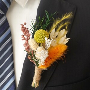 May include: A colorful boutonniere featuring a cluster of dried flowers in shades of yellow, orange, white, and pink, accented with green foliage. The stems are wrapped with twine. The boutonniere is pinned to a black suit jacket.