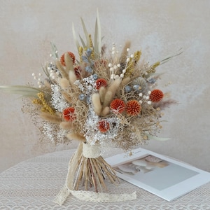 Könnte beinhalten: Ein Trockenblumenstrauß mit orangefarbenen, weißen und gelben Blüten, mit beige- und grüntönen. Der Strauß ist mit einem beigen Spitzenband gebunden und liegt auf einer weißen Spitzentischdecke.