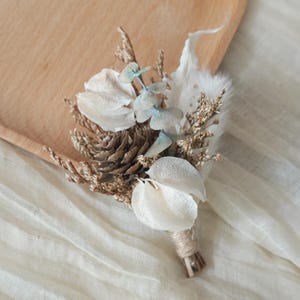 Könnte beinhalten: Handgefertigte Boutonniere mit getrockneten Blumen und Blättern. Die Anordnung umfasst einen Tannenzapfen, weiße Blütenblätter und Zweige aus blau-grünem Eukalyptus. Die Stiele sind mit Bindfaden umwickelt.