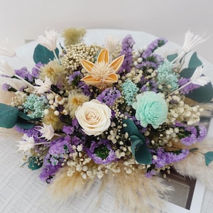 Könnte beinhalten: Ein lebendiges Blumenarrangement mit einer cremefarbenen Rose, lila Statice und hellblauen Akzenten. Der Strauß enthält getrocknetes Pampasgras, Eukalyptusblätter und andere Trockenblumen, wodurch eine strukturierte und farbenfrohe Darstellung entsteht.