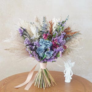 Könnte beinhalten: Ein Strauß Trockenblumen mit blauen Rosen, lila Strandflieder und Pampasgras. Die Stiele sind mit einem beigen Band gebunden. Eine kleine weiße Engelsfigur steht neben dem Strauß auf einem Holztisch.