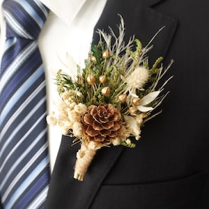 May include: A rustic boutonniere featuring a brown pinecone, dried flowers, and greenery. The arrangement includes gold accents and is wrapped with twine. The boutonniere is pinned to a black suit jacket, next to a blue and white striped tie.