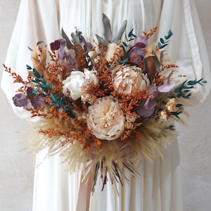 Può includere: Un bouquet di fiori secchi con una varietà di trame e colori. L'arrangiamento include grandi peonie bianco crema, accenti color ruggine, fogliame turchese e tocchi di viola e beige. Il bouquet è legato con un nastro marrone chiaro.