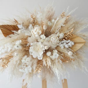 Könnte beinhalten: Ein dekoratives Blumenarrangement mit flauschigem beigem Pampasgras, weißen Trockenblumen und goldfarbenen Palmblättern. Die Komposition enthält runde weiße Samenkapseln und zartes Blattwerk, was für eine strukturierte Optik sorgt.