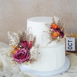 Könnte beinhalten: Zweistöckige weiße Torte, verziert mit weinroten und orangenen Blumen, Pampasgras und kleinen weißen Perlen. Der Kuchen steht auf einem weißen Kuchenständer. Das Wort "OCT" ist auf einem Holzschild im Hintergrund zu sehen.