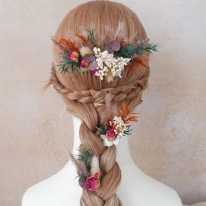 May include: A braided hairstyle decorated with floral hair accessories. The hairpieces feature dried flowers in shades of pink, orange, white, and green, with feathery plumes. The hair is a light brown colour.