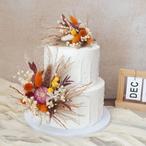 Könnte beinhalten: Ein zweistöckiger weißer Kuchen, verziert mit Trockenblumen in Orange-, Gelb- und Burgundertönen. Der Kuchen steht auf einem weißen Kuchenständer. Ein Holz-Kalender zeigt "DEC 02".