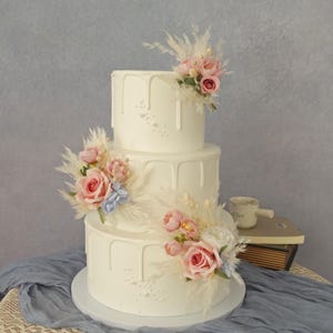 Könnte beinhalten: Ein dreistöckiger weißer Kuchen mit Tropfendesign, verziert mit rosa Rosen, blauen Blumen und federartigen Akzenten. Der Kuchen steht auf einer weißen Basis, ideal für eine Hochzeit. Der Kuchen ist mit einem leichten weißen Sprühnebel verziert.