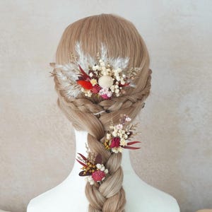 Könnte beinhalten: Geflochtene Frisur mit drei floralen Haaraccessoires. Die Haarstücke zeigen eine Mischung aus getrockneten Blumen in Rot, Weiß und Creme, mit Akzenten aus federartigen Federn. Die Accessoires sind am Oberkopf und entlang des Zopfes platziert.