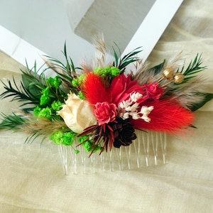 Könnte beinhalten: Ein Haarkamm mit einem floralen Arrangement aus roten, weißen und grünen Blüten, ergänzt durch grüne Zweige und goldfarbene Perlen. Der Kamm besteht aus transparentem Kunststoff und wird ins Haar gesteckt.