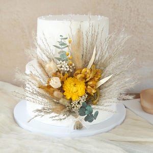 Könnte beinhalten: Zweistöckige weiße Torte, verziert mit Trockenblumen und Weizen. Der Kuchen ist mit einer großen gelben Blume, Eukalyptus und anderen getrockneten floralen Elementen geschmückt. Der Kuchen steht auf einem weißen Kuchenständer.