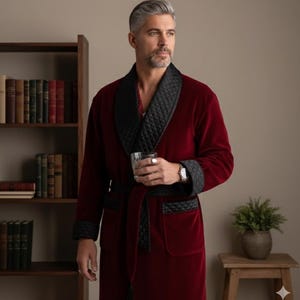 May include: A burgundy velvet robe with a black quilted shawl collar, cuffs, and belt. The robe has pockets and a matching belt. The person is holding a glass of water. The robe is a luxurious garment.