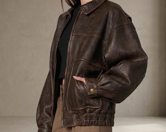 May include: A dark brown leather bomber jacket featuring a classic collar, long sleeves with cuffs, a front zipper closure, and two large patch pockets on the front with snap button details. The jacket has an elasticized hemline.