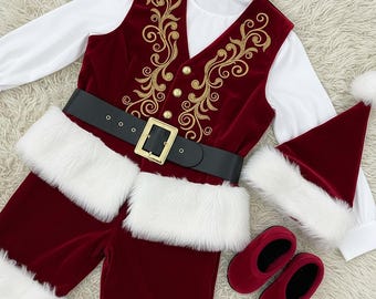 Könnte beinhalten: Ein Weihnachtsmann-Kostüm-Set mit einer bordeauxroten Samtweste mit goldener Stickerei, einem schwarzen Gürtel mit goldener Schnalle, einer passenden Hose mit weißem Kunstfellbesatz, einem Hut, einem weißen Langarmshirt und roten Samtslippers. Das Outfit ist für einen festlichen Look.