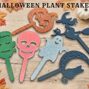 May include: Six Halloween plant stakes featuring a glittery orange pumpkin, a black witch on a broomstick with a cat, a green Frankenstein monster, a pink skull, a silver glittery ghost, and a black crescent moon with a cat silhouette.
