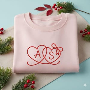 May include: A light pink sweatshirt with a red embroidered design of two intertwined hearts, the letters "A" and "S" are inside the hearts, and a bow. The sweatshirt is folded neatly on a light beige surface.