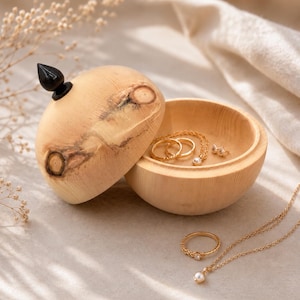 Può includere: Un portagioie rotondo in legno chiaro con coperchio rimovibile dotato di un pomello scuro e appuntito. La scatola presenta venature e nodi del legno visibili. All'interno del contenitore aperto ci sono anelli e una collana dorati, con altri gioielli.