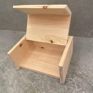 Könnte beinhalten: Eine helle, unbehandelte Holzkiste mit einem aufgeklappten Scharnierdeckel. Die Kiste weist eine sichtbare Holzmaserung und natürliche Astlöcher auf, mit einem rechteckigen Boden und zwei Seitenwänden, die den Deckel in der offenen Position stützen.