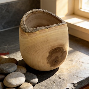 May include: A handcrafted wooden vase with a natural, organic shape. The light-colored wood features visible grain and a dark knot. The rim has a textured, bark-like edge. A collection of smooth, gray stones rests nearby.