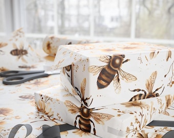 May include: Two wrapped gift boxes with bee and floral print. The boxes are stacked on a table covered with matching wrapping paper. A roll of paper, scissors, and a gray ribbon are also visible.