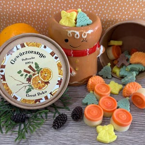 May include: A seasonal display with a gingerbread man-shaped container holding tree and bow-shaped wax melts. A round container with the text "Gewürzorange" and a whole orange are also present. The scene is decorated with pinecones and greenery.