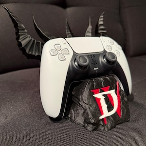 Könnte beinhalten: Ein weiß-schwarzer PlayStation 5-Controller auf einem individuellen Ständer. Der Ständer ist ein schwarzes Felsdesign mit den Buchstaben "D" und "W" in Rot und Weiß. Schwarze Hörner erstrecken sich nach oben.