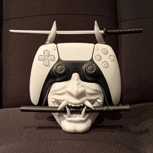 May include: A white and black PlayStation 5 controller rests on a white, textured Oni mask. Two small white swords are positioned above the controller, and a black sword is held in the mask's mouth. The design is a fusion of gaming and Japanese culture.