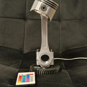May include: A silver-colored piston lamp with a black gear base. The lamp has a remote control with multiple color options. The lamp is made of metal and has a unique industrial design.