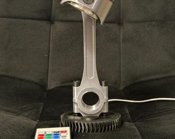 Op de afbeelding: Een zilverkleurige zuigerlamp met een zwarte tandwielbasis. De lamp heeft een afstandsbediening met meerdere kleuropties. De lamp is gemaakt van metaal en heeft een uniek industrieel ontwerp.